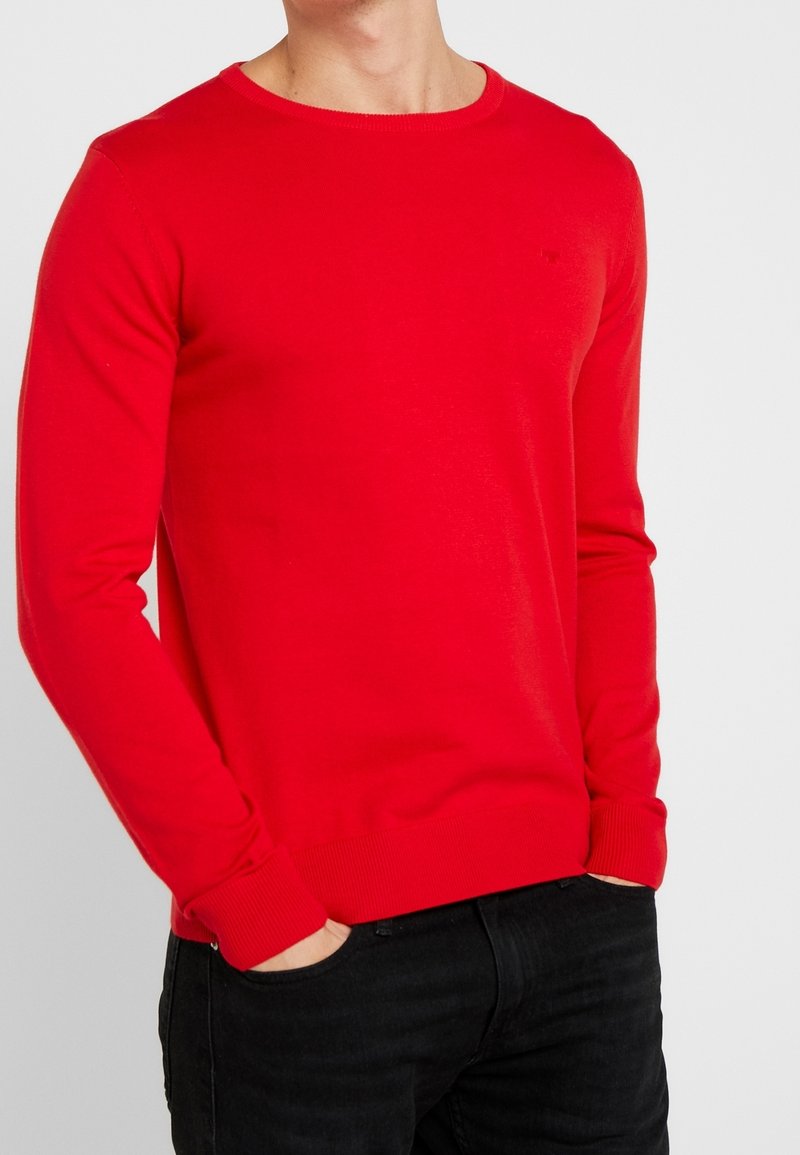 Pull en tricot rouge avec un col rond, des manches longues et des poignets côtelés. Porté avec un jean foncé, présentant un détail de logo subtil sur la poitrine.