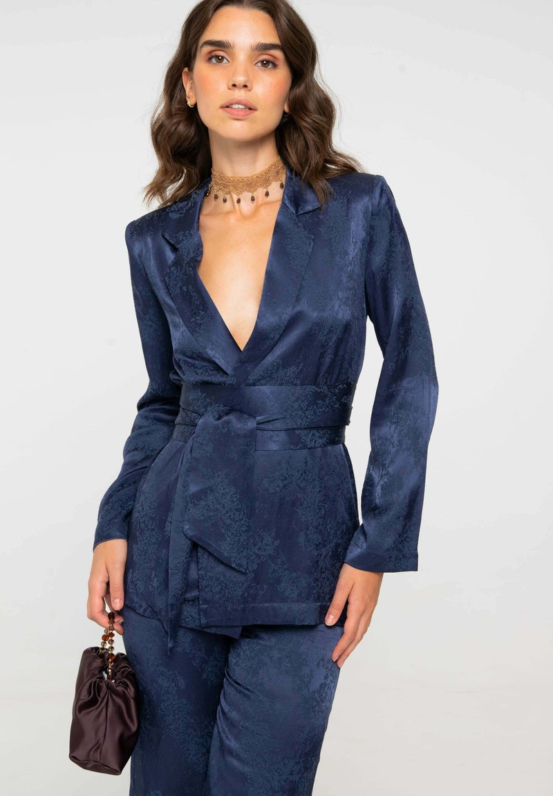 Mujer con un traje de satén azul profundo con estampado, cinturón ancho, sosteniendo un pequeño bolso de satén con asa de cuentas y un collar de encaje tipo choker.