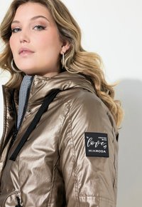 Chaqueta de bronce metálico con cierre de cremallera, capucha y detalles en negro. Incluye una etiqueta con el texto "Creada con amor" adjunta a la manga.