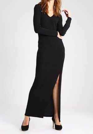 Kobieta w długiej czarnej sukience z dekoltem w kształcie litery V, z długimi rękawami i rozcięciem do uda, w czarnych butach na wysokim obcasie.