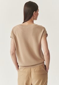 Beige gebreid T-shirt met korte mouwen en een ribgebreide halslijn en zoom, gecombineerd met lichtbruine nauwsluitende broek met achterzakken.