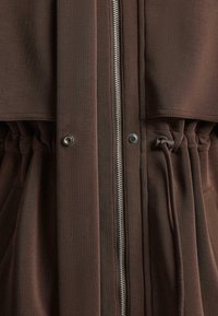 Brown jacket featuring a zipper closure, elastic drawstring at the waist, and snap button details. Smooth texture with a relaxed fit design.