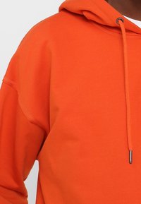 Gros plan sur un sweat à capuche orange avec cordons de serrage et embouts métalliques, porté par une personne sur un fond uni.
