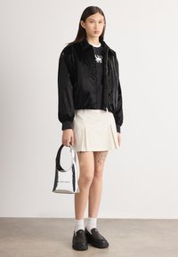 Jachetă bomber lucioasă de culoare neagră peste un tricou alb cu grafică, fustă plisată de culoare bej, pantofi negri slip-on și poșetă argintie cu logo.