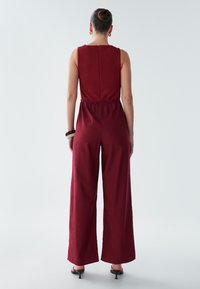 Bordo kombinezon z nazaj z nabranim detajlom, širokimi hlačnicami, elastičnim pasom in zgornjim delom brez rokavov. Tekstura je gladka in lahka.
