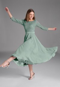 Robe verte menthe avec un corsage en dentelle présentant des motifs complexes et des paillettes ; jupe en chiffon fluide ajoute du mouvement. Des talons argentés complètent le look.