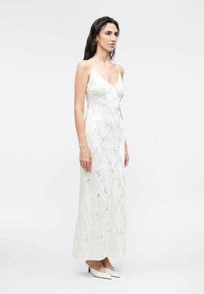 Frau trägt ein langes weißes Blumenbesticktes Trägerkleid mit dünnen Trägern und weißen spitzen Schuhen, stehend vor einfachem Hintergrund.