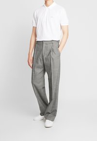 Polo blanc avec un petit logo, associé à un pantalon gris clair à carreaux avec des plis. Le mannequin porte des baskets blanches.