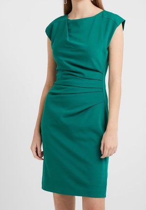 Kobieta ubrana w turkusową sukienkę bez rękawów, sięgającą do kolan, z marszczeniami z przodu i delikatnymi poduszkami na ramionach, na jednolitym tle.