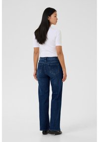 Pikajuukseline mustade juustega naine seisab selg vaataja poole, seljas valge lühikeste varrukatega ülaosa, tumelillad teksapüksid ja mustad kingad, lihtsa taustaga.