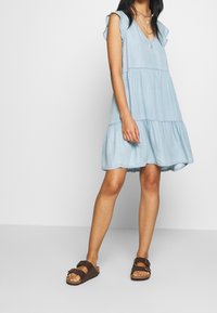 Vaaleansininen denim-mekko, jossa on lyhyet röyhelöhihat, V-kaula-aukko, kerroksellinenhelma ja rento istuvuus, yhdistettynä ruskeisiin sandaaleihin.