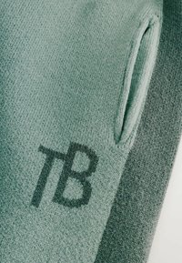 Švelnaus žalio spalvos trikotažas su nykščio anga, tamsiai pilkų akcentų ir įspaustu "TB" logotipu arti apatinės dalies. Tekstūra atrodo lygi.