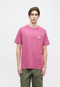 Růžové bavlněné tričko s krátkým rukávem a kulatým výstřihem. Na levé straně hrudi má bílé logo. Kombinováno s olivově zelenými kalhotami.