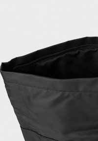 Sac fourre-tout en tissu noir avec une forme structurée, présentant une texture lisse et une ouverture supérieure. La doublure intérieure est également noire.