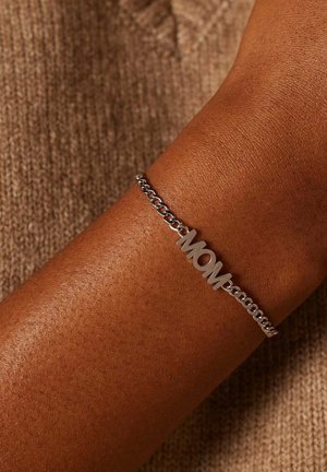 Zilveren schakelarmband met "MOM" gegraveerd. Bevat een gladde textuur en een eenvoudig, modern ontwerp. Draagt op een gebruinde huid.