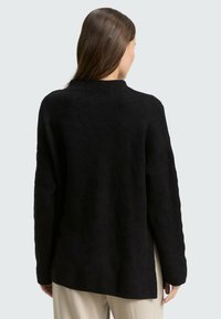 Vrouw met lang bruin haar, gekleed in een losse zwarte textuursweater en beige broek, gezien van achteren tegen een effen lichte achtergrond.