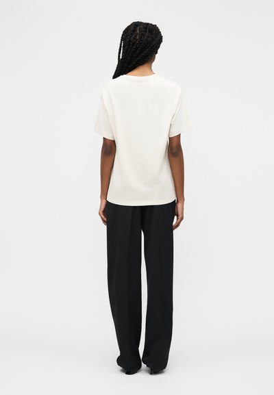 Άτομο με μακριά πλεγμένα μαλλιά, φαινόμενο από πίσω, φορώντας κοντομάνικο λευκό πουκάμισο και φαρδιά μαύρη παντελόνα σε απλό φόντο.