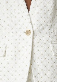 Veste en tweed blanc avec un motif matelassé, des accents dorés et un détail de bouton rond en or. Présente une surface texturée prononcée avec une légère brillance.