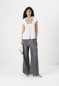 Blouse blanche avec broderie florale sur le col, associée à un pantalon large gris taille haute. Un sac à main et des chaussures argentés complètent le look.