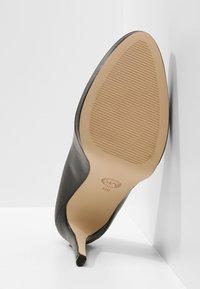 Chaussure à talon haut en cuir noir positionnée semelle vers le haut montrant la semelle texturée couleur tabac et un petit talon aiguille sur fond blanc.
