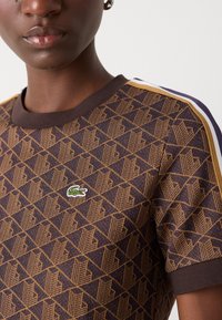 Braunes T-Shirt mit geometrischem Muster, geripptem Rundhalsausschnitt und Schulterstreifen in Creme und Lila. Mit kleinem grünen Logo.