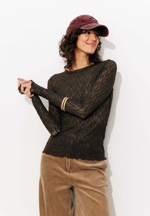 Femme portant un haut transparent à manches longues noir, un pantalon en velours côtelé beige, une casquette violette et des bracelets en or, souriant et joignant les mains.