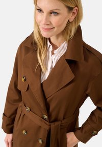 Cappotto lungo marrone con colletto ampio, bottoni doppi e cintura coordinata. Tessuto liscio con una leggera lucentezza. Camicia a righe sottostante.