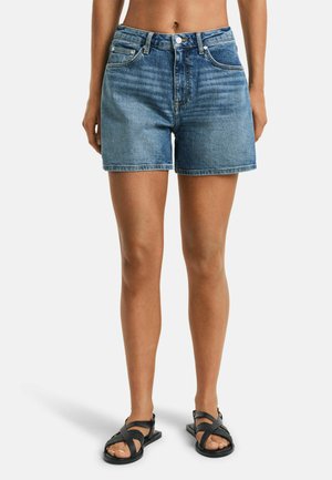 Helle blaue Denim-Shorts mit klassischem Fünf-Taschen-Design, Knopfverschluss und rohem Saum. Getragen mit schwarzen Sandalen mit Riemen.