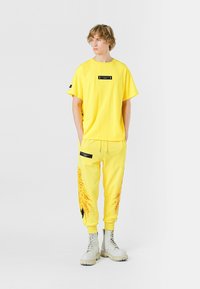 Camiseta de algodón amarilla con un parche negro y joggers a juego con un diseño impreso. Usada con botas de combate grises con cordones.