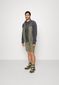 Fleecejakke i grå og grøn, med høj krave, lynlås foran, båret over en sort t-shirt og olivenfarvede shorts, parret med mønstrede sneakers.