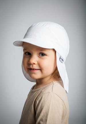 Young child wearing a white sun hat with neck flap and a beige shirt, smiling gently against a plain gray background.