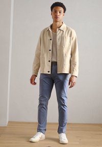 Beżowa bawełniana kurtka z czarnymi guzikami, jasnoszary sweter typu knit, niebieskie spodnie o slim fit i białe sneakersy z delikatnymi beżowymi akcentami.