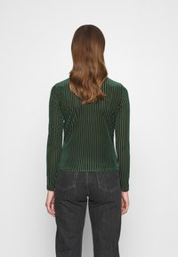 Grünes, geripptes langärmliges Oberteil aus Samt mit taillierter Silhouette und Rundhalsausschnitt, kombiniert mit schwarzen Jeans.