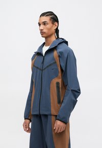 Αδιάβροχο μπουφάν με κουκούλα, που διαθέτει μπλε άνω τμήμα και καφέ κάτω τμήμα. Περιλαμβάνει τσέπη με φερμουάρ στο πλάι και μαύρες λεπτομέρειες.