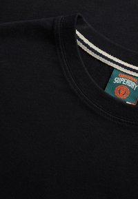 Maglietta nera a girocollo con cuciture dettagliate e un'etichetta verde ben visibile con il logo "Superdry" in arancione e bianco.