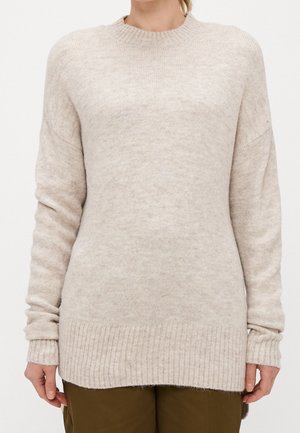 Frau trägt einen beigefarbenen langärmligen Strickpullover mit gerippten Bündchen und Saum, kombiniert mit olivgrünen Hosen, steht vor einem schlichten Hintergrund.