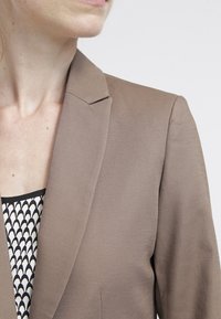 Hnědý přizpůsobený blejzr s hladkou texturou, jednobuttonovým zapínáním a klopovými límci, nošený přes vzorovaný top s černobílými šupinami.