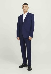 Granatowy garnitur wykonany z teksturowanej tkaniny, z zapięciem na jeden guzik i klapami w kształcie litery Notch. Połączony z białą koszulą i czarnymi butami.