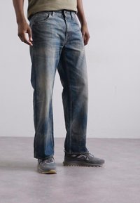 Jeans en denim avec une coupe droite classique, présentant un délavé bleu clair, une texture usée et un design à cinq poches, portés avec des chaussures de sport grises.