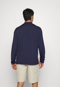 En man som står vänd bortåt, iförd en långärmad marinblå skjorta och beige knälånga shorts mot en enfärgad vit bakgrund.