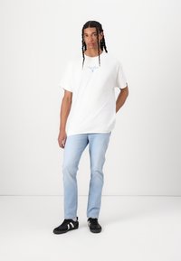 Witte katoenen t-shirt met blauwe grafische details op de borst, gecombineerd met lichtblauwe spijkerbroeken en zwarte sneakers met witte strepen.