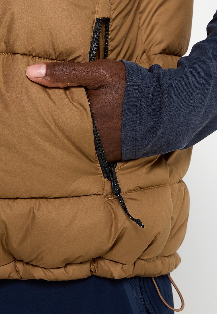 Brązowa kamizelka puchowa z pikowanym wzorem, z zamykaną na zamek kieszenią oraz teksturowanym materiałem. Koszula pod spodem jest w kolorze granatowym.