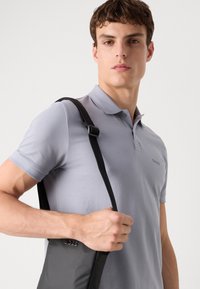 Polo de color gris claro hecho de algodón, con un cuello de tres botones y logo bordado, combinado con un bolso negro con correa para el hombro.