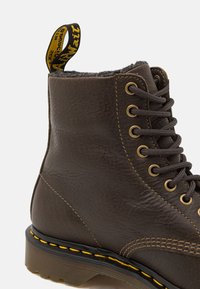 Botte en cuir marron à lacets avec coutures jaunes, semelle épaisse, et une languette arrière noire et jaune avec logo.
