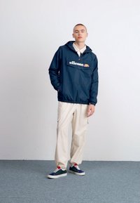 Giacca a vento marina con tasca frontale e logo "ellesse", abbinata a pantaloni cargo color beige chiaro e sneaker navy con lacci multicolori.