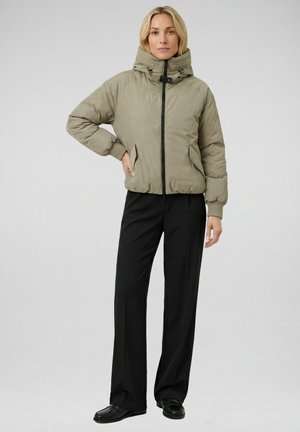 Vrouw draagt een licht olijfgroene jas met hoge kraag, zwarte broek met wijde pijpen en zwarte schoenen, staand tegen een effen achtergrond.