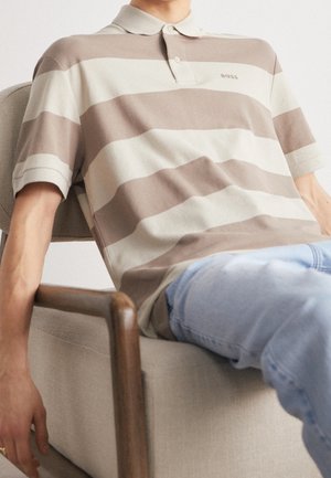Person, die auf einem beigen Stuhl entspannt sitzt, trägt ein braun-weiß breit gestreiftes Poloshirt und hellblaue Jeans, mit dem Arm auf der Armlehne des Stuhls ruhend.