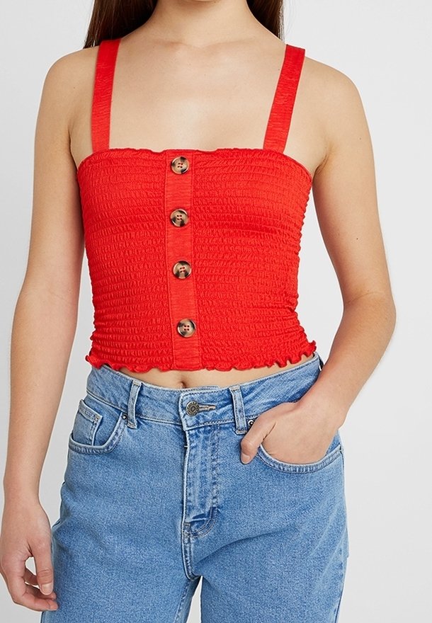 Top corto rojo con textura fruncida, tirantes anchos y cinco botones de madera en la parte delantera. Combinado con jeans azul de cintura alta.
