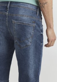 Jeans od trapera u tamnoplavoj boji, s uskim krojem, vidljivim šavovima i stražnjim džepom s karakterističnim V-oblikovanim detaljem.
