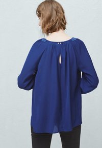 Blusa blu navy con maniche lunghe, tessuto morbido, scollatura arricciata e dettaglio sul retro con chiusura a chiave e due bottoni al colletto.